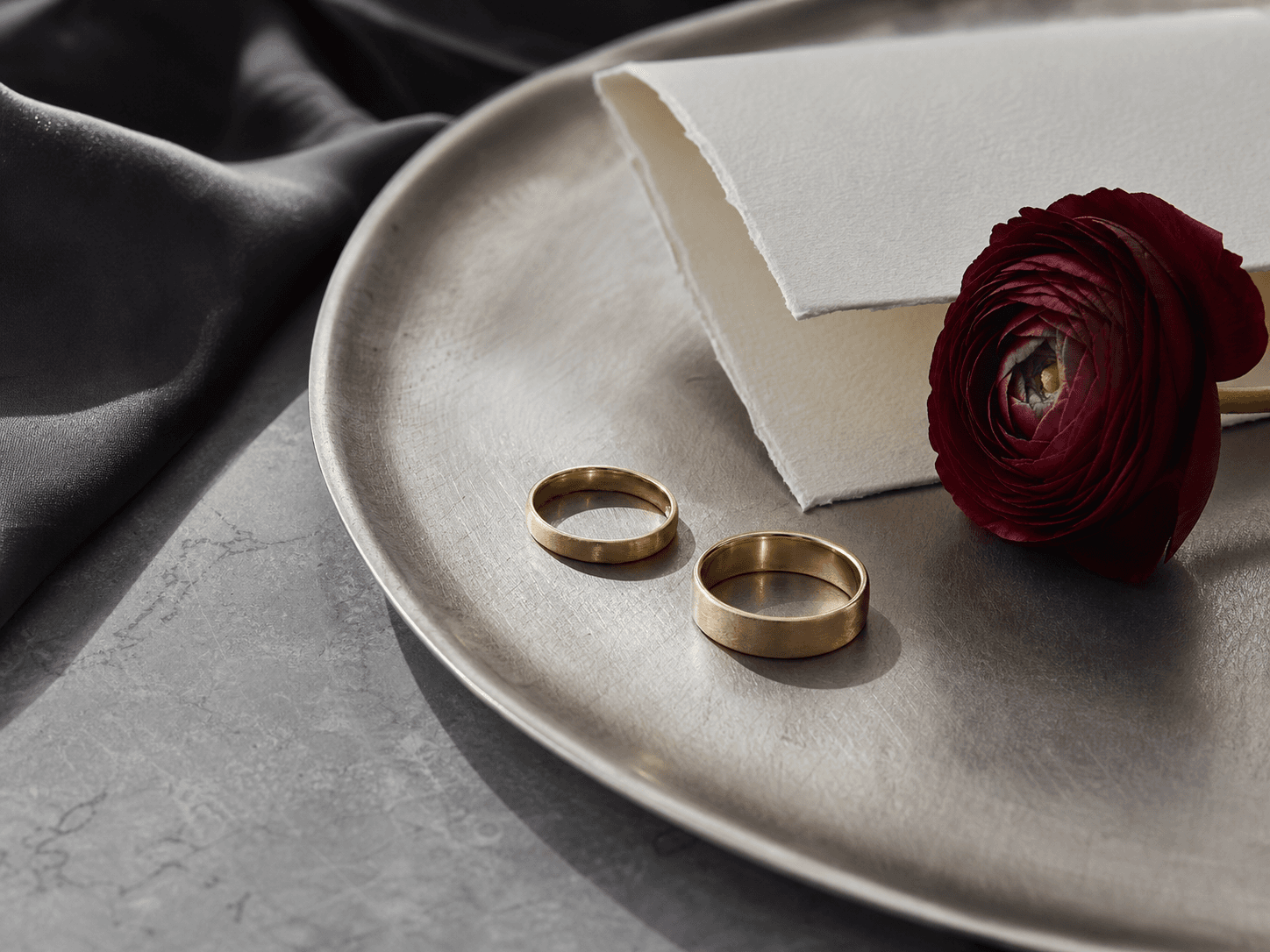Two gold wedding bands beside a deep red flower and vow paper